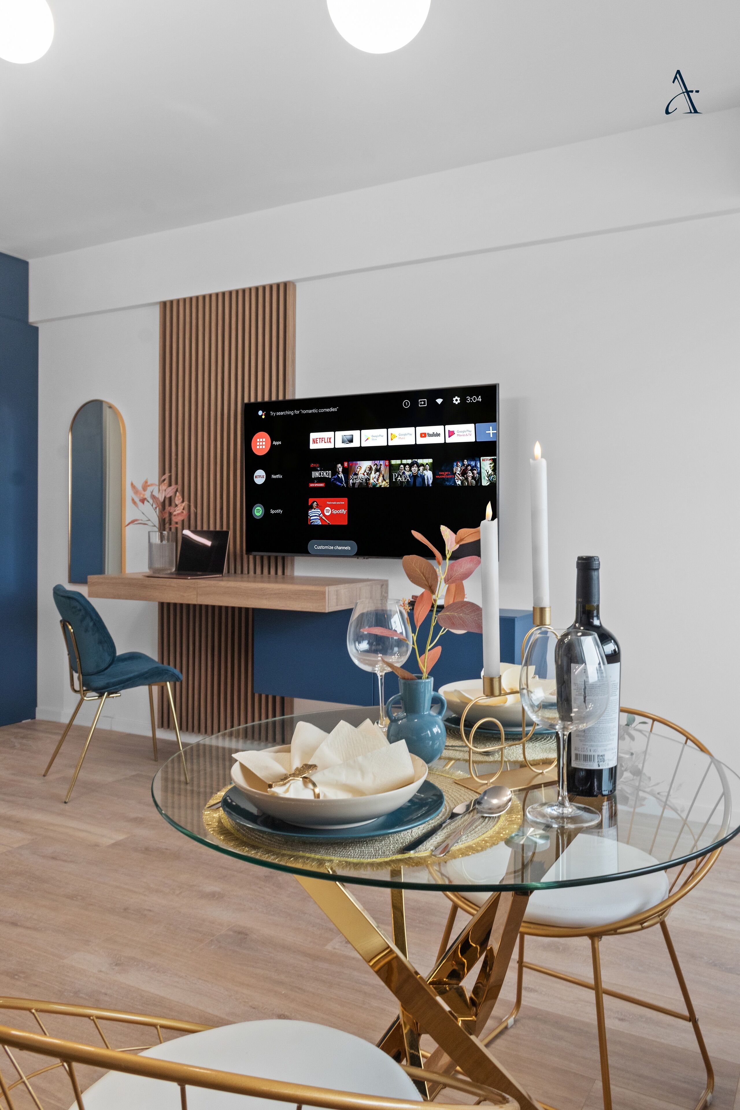 A modern dining area with a glass table, gold chairs, and a flat-screen TV mounted on the wall displaying streaming services.