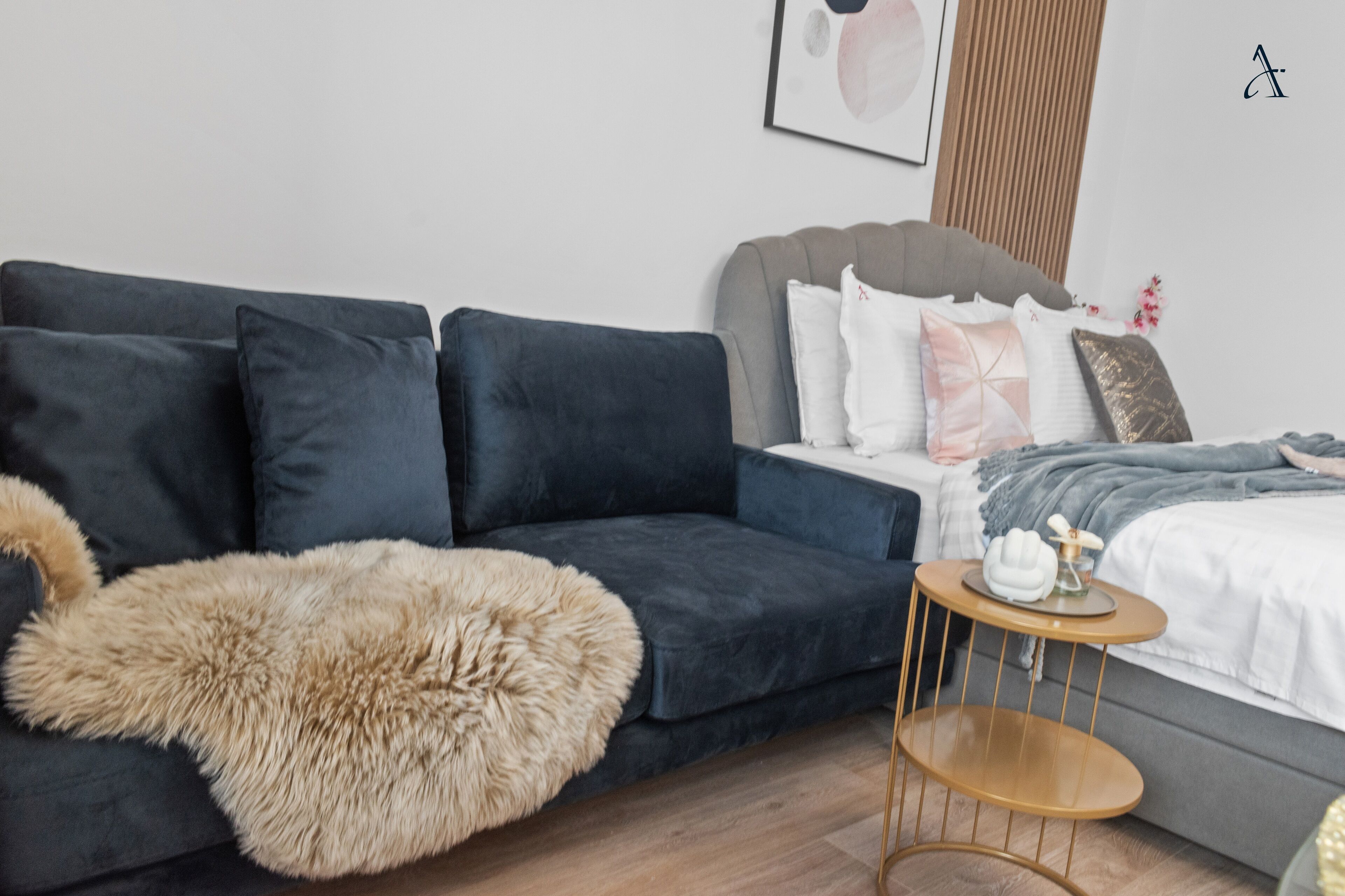 Un dormitorio moderno con un sofá azul oscuro, un plaid beige suave, una pequeña mesita de noche de madera y una cama con muchas almohadas.