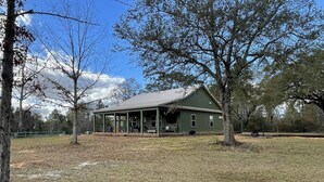 A green building with a porch, surrounded by trees and open grassy area.