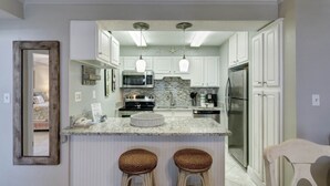 Una cocina moderna con armarios blancos, encimeras de granito y electrodomésticos de acero inoxidable. Hay dos taburetes de bar y lámparas colgantes sobre la isla.