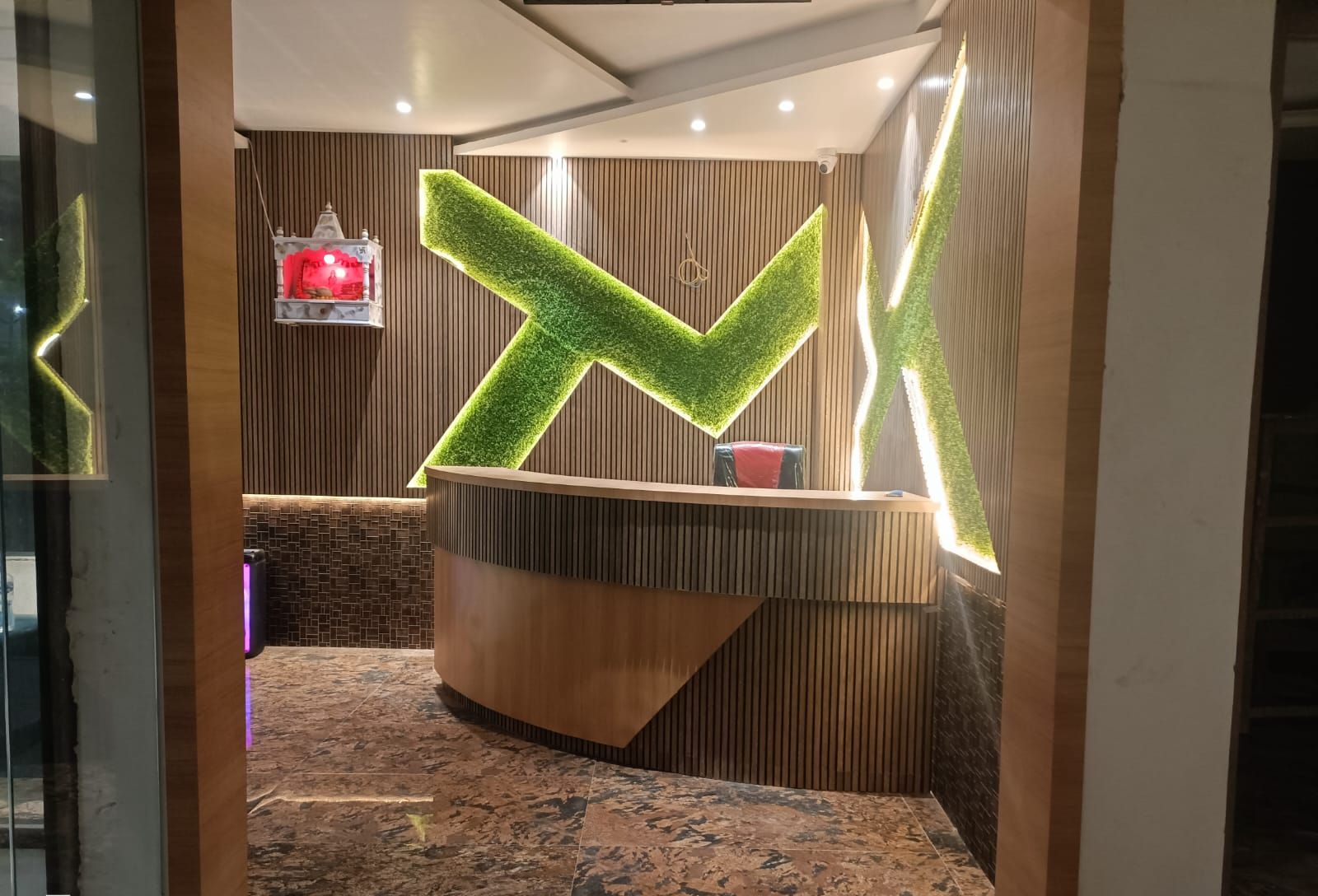 Zona de recepción con mostrador de madera, un letrero iluminado verde en forma de 'X' y un objeto rojo pequeño en la pared.