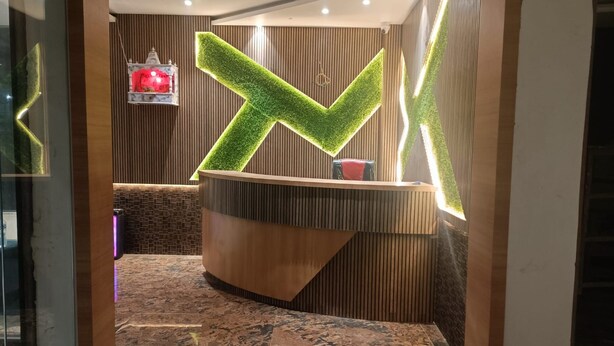 Una zona d'entrada amb un taulell de recepció de fusta, un senyal il·luminat verd en forma de 'X' i un petit objecte vermell a la paret.