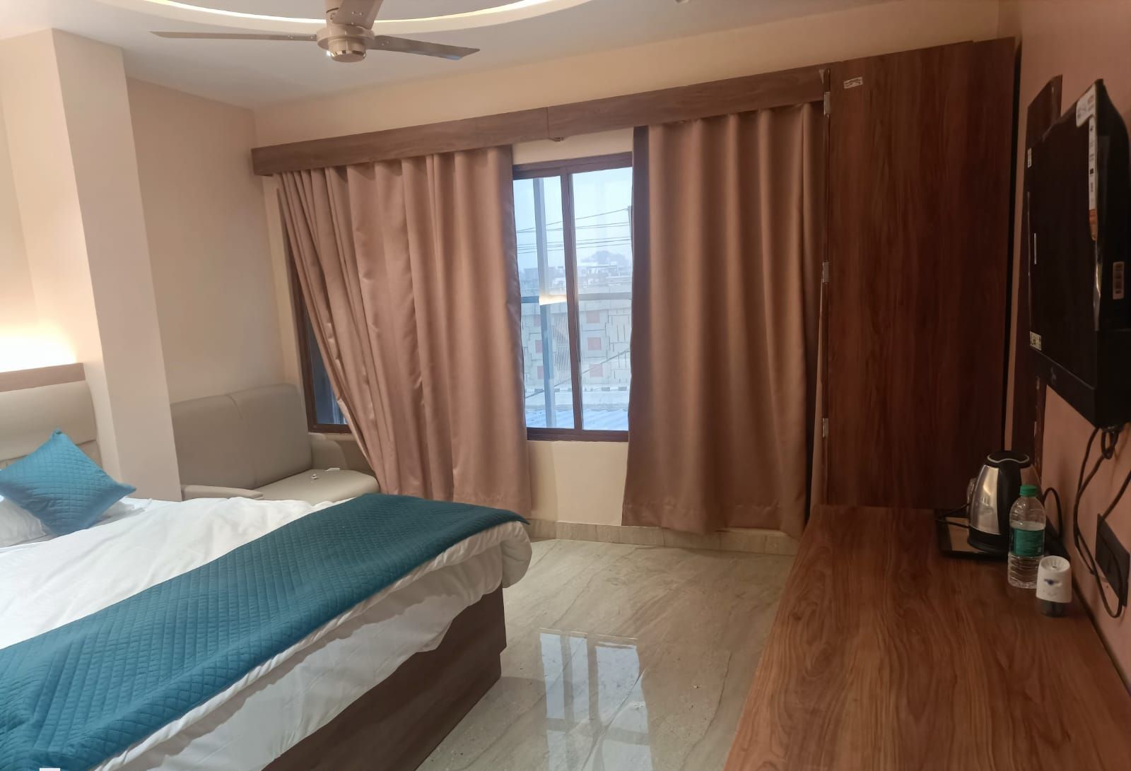Ein Hotelzimmer mit Bett, Schreibtisch, Stuhl, Deckenventilator, Fenster mit Vorhängen und Fernseher.
