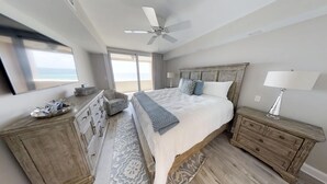A bedroom with a large bed, a ceiling fan, a nightstand with a lamp, and a dresser.