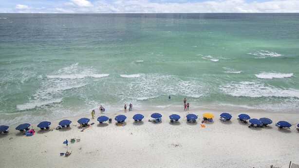 Een strand met helder turquoise water, een zandstrand en diverse blauwe parasols.