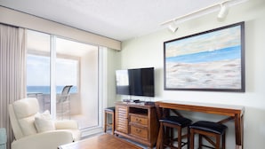 Ein Hotelzimmer mit großem Fenster, Flachbildfernseher, Holztisch, zwei Barhocker, weißem Sessel und einem Gemälde mit Strandmotiv.