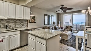 A modern kitchen with white cabinets, granite countertops, and a large island. There is a dining area with a white table and chairs, and a balcony with a view of the ocean. - Sunset Jetty View! Enjoy one free beach chair set up and umbrella each day! (Destin)