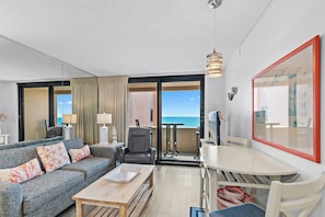 A modern living room with a sofa, coffee table, and dining table, a balcony with a view of the sea, and a large framed artwork on the wall.