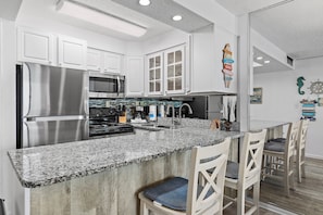 A kitchen with white cabinets, stainless steel appliances, a granite countertop, and a bar area with white chairs.