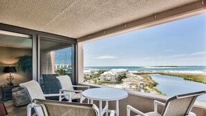 A balcony with white wicker chairs and a small round table, overlooking a beach and a body of water with buildings in the background.