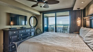 A bedroom with a large bed, a dresser, a flat-screen TV, a ceiling fan, and a view of the beach through the window.