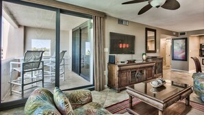 A spacious living room with a large sliding glass door leading to a balcony, a ceiling fan, a flat-screen TV, and a wooden cabinet.