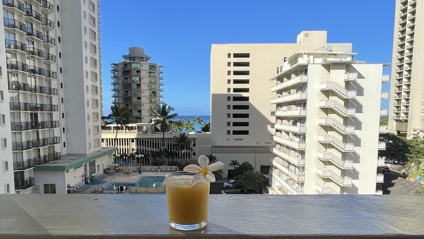 Un balcon où l’on peut prendre un verre, avec vue sur une piscine et des immeubles de grande hauteur, sous un ciel bleu dégagé.