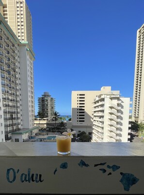 A balcony with a drink, overlooking a pool and high-rise buildings, with a clear blue sky. - Contemporary Ocean View Suite - In the Heart of Waikiki (Honolulu)