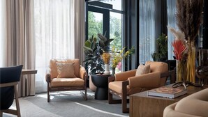 A modern living room with two leather sofas, a coffee table, and a side table. There is a large window with sheer curtains, and a potted plant with yellow flowers. - Ada Hotel (Ada)