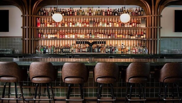A bar area with a wooden ceiling, multiple shelves of liquor bottles, and four bar stools. - Ada Hotel (Ada)