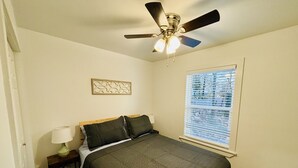 Ein Schlafzimmer mit Bett, Nachttisch, Lampe, Deckenventilator, Fenster mit Jalousien und einer Wanddekoration.
