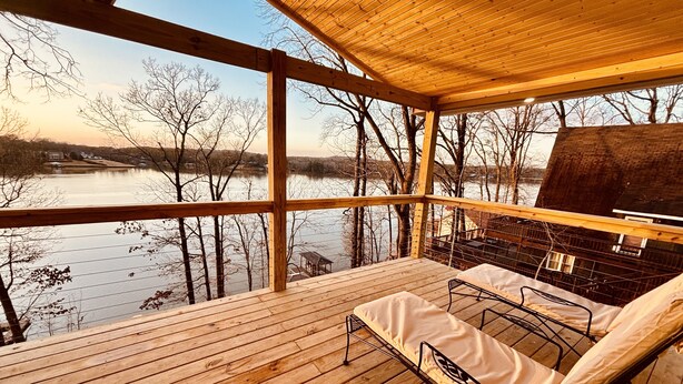 Eine Holzterrasse mit Blick auf einen See und Bäume, ein Grill und eine Metallbank mit Kissen.