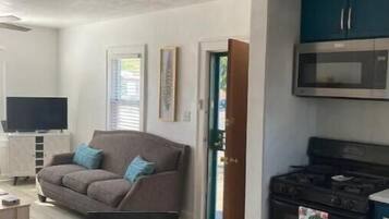 A living room with a sofa, a coffee table with snacks, a TV, and a kitchen area with a stove and microwave.