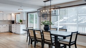 Een moderne keuken met witte kasten, een eetgedeelte met een donkere tafel en rieten stoelen, en een groot raam met zonwering.