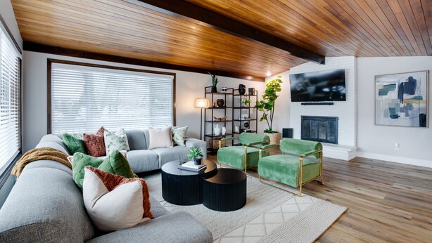 Een moderne woonkamer met een houten plafond, een grijze bank, groene fauteuils, een zwarte salontafel en een boekenkast.