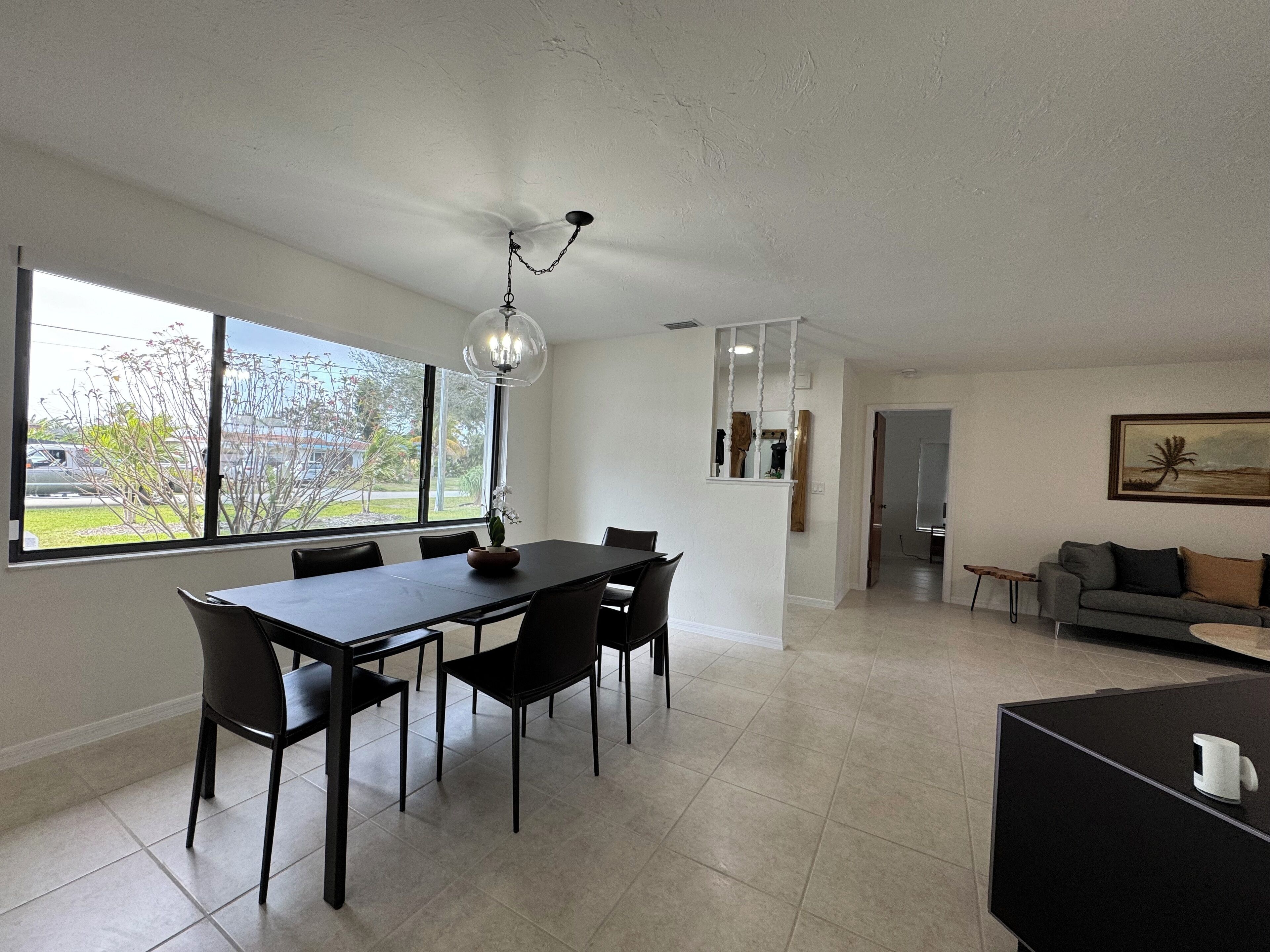 A modern living room with a dining area, large windows, a ceiling fan, and a painting on the wall.