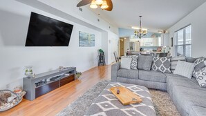 Een moderne woonkamer met een grijze bank, houten vloer, plafondventilator, flatscreen televisie en een keuken op de achtergrond.