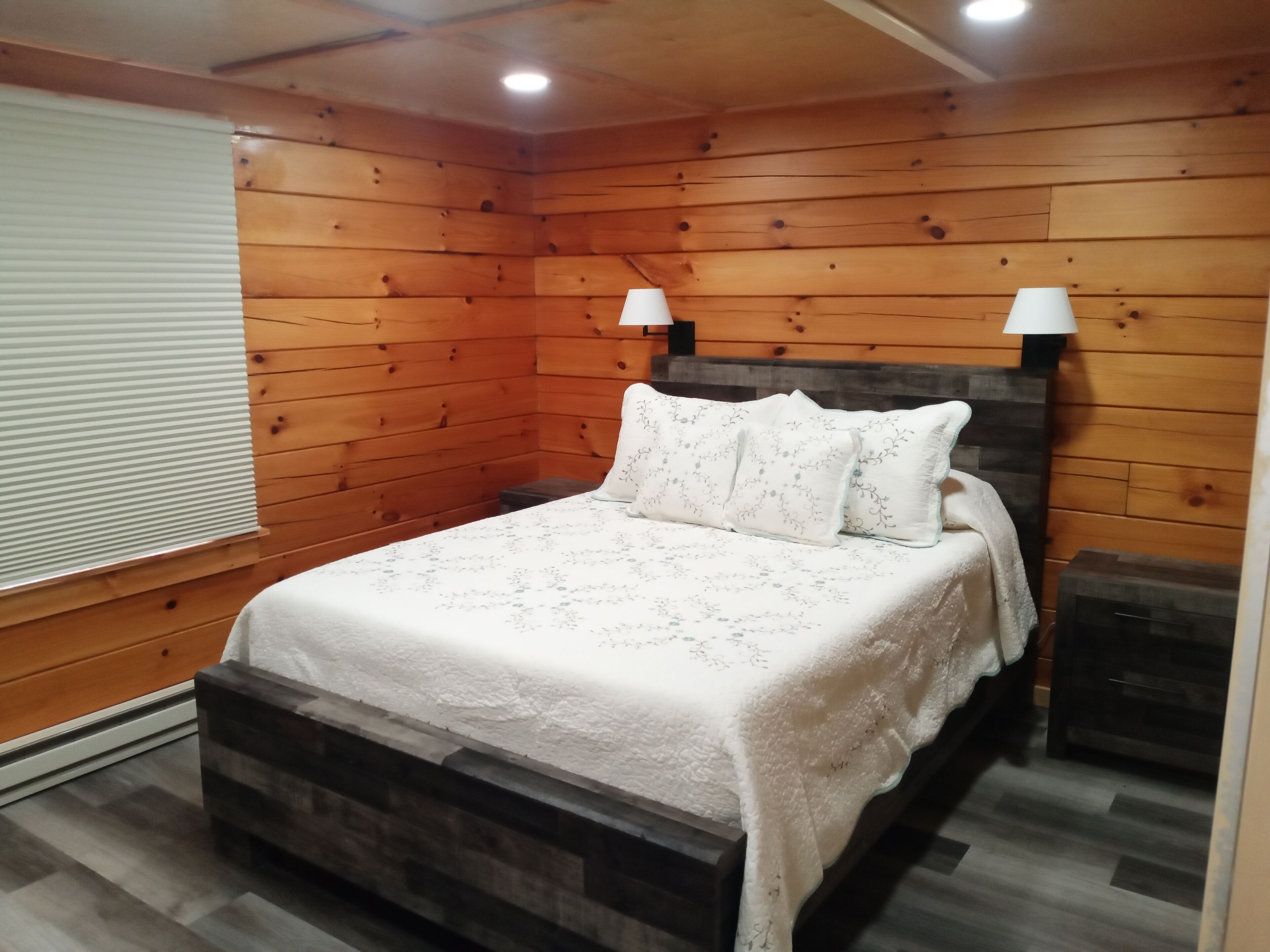Een slaapkamer met houten lambrisering, een bed, twee wandlampen en een raam met zonwering.