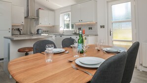 A modern kitchen with white cabinets, a wooden dining table set for two, and a large window providing natural light. - Buttercup Lodge (Dartmouth)