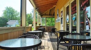 A covered outdoor seating area with tables and chairs, large windows, and a view of greenery.