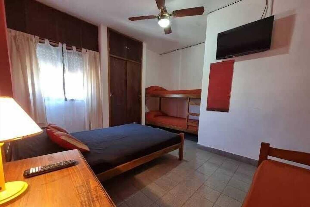 A room with a bed, a wooden table with a lamp, a ceiling fan, and a television mounted on the wall.