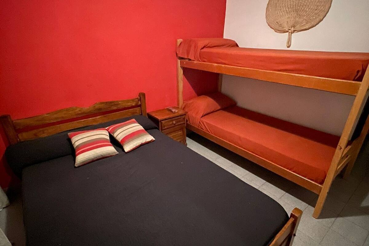 A bunk bed with a black mattress and two pillows, a wooden nightstand with a book, and a red wall with a woven basket hanging.
