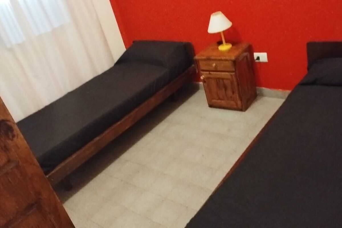 A room with two single beds, a small wooden nightstand with a lamp, and a white curtain.