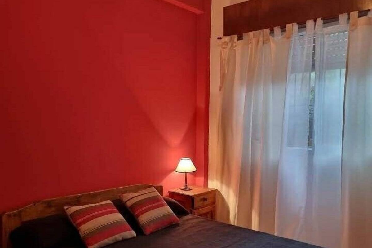A single bed with a wooden headboard, a bedside table with a lamp, and a window with sheer curtains.