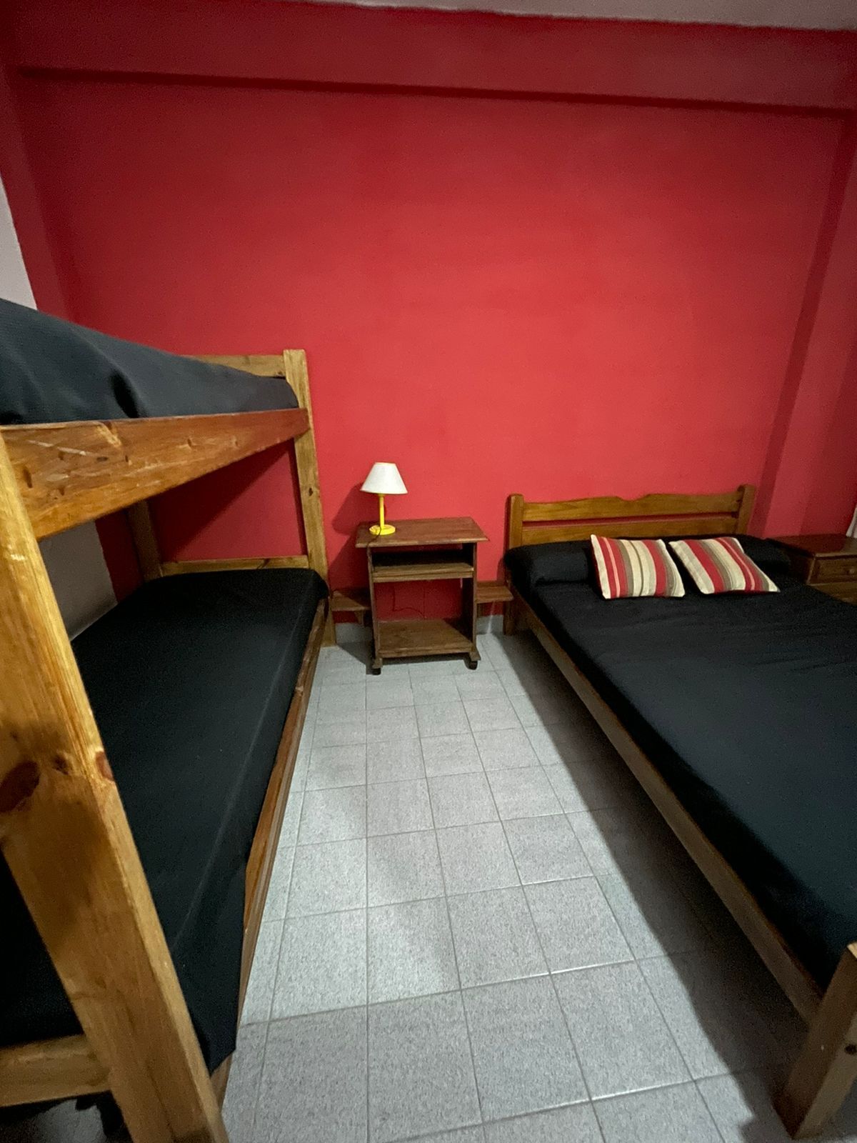 Una habitación pequeña y funcional con dos literas y una cama individual, una mesita de noche de madera con lámpara, y una pared roja.