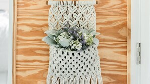 Een macramé wanddecoratie met een vogelillustratie en een lijst er boven.