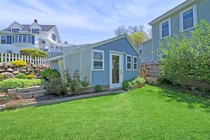 A residential property with a well-maintained lawn, a wooden fence, and a small garden area with various plants. - 2BR/2BA Harborside Home · Fenced Backyard · Ocean View ·Walk to Rocky Neck+Beach (Gloucester)