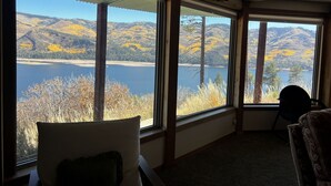 Una habitación con amplios ventanales que ofrecen vistas a un lago y colinas. Hay dos sillones y un radiador.
