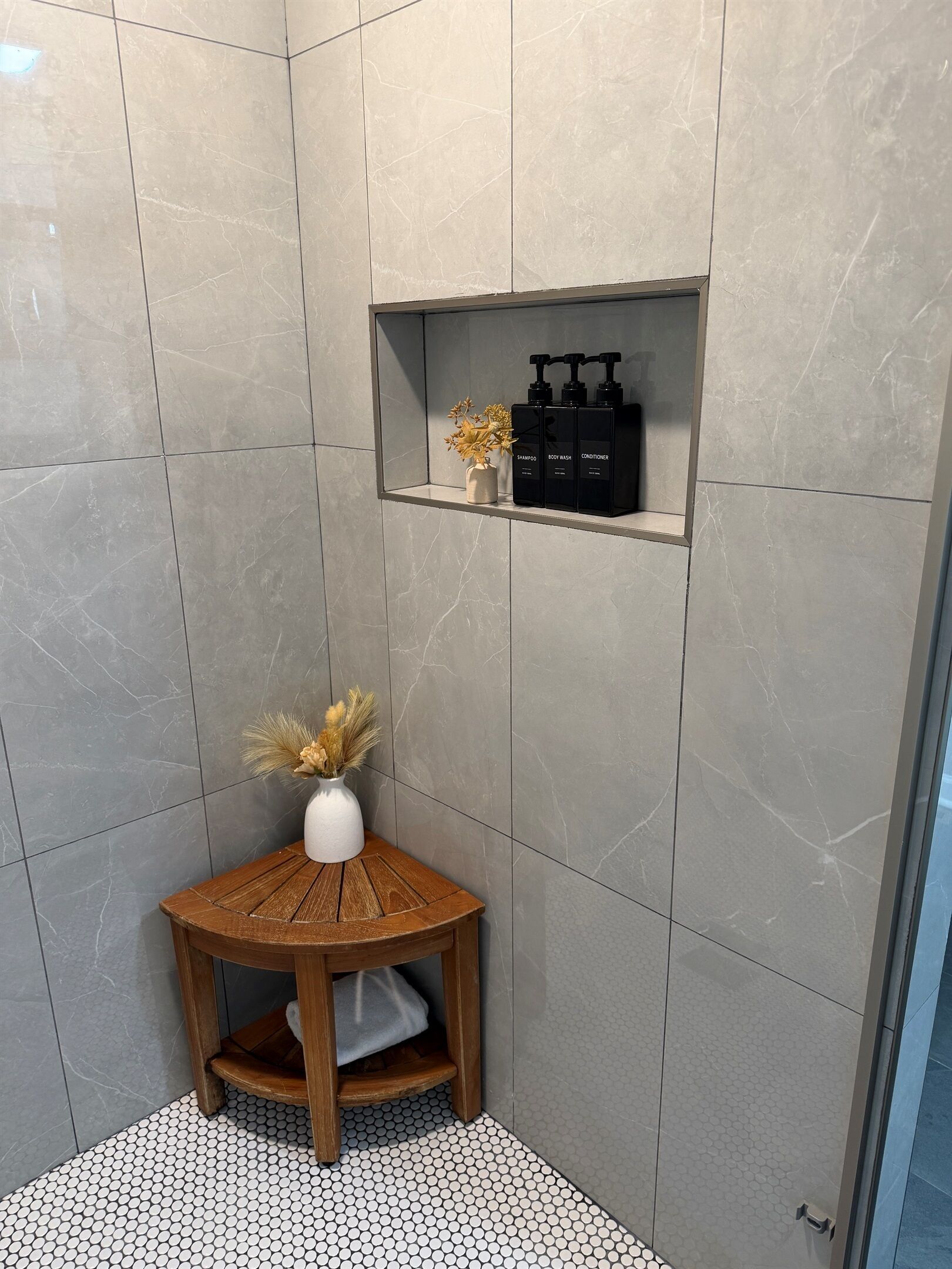 Un baño con suelo de baldosas, un taburete de madera con un florero de flores secas y un estante con artículos de aseo.