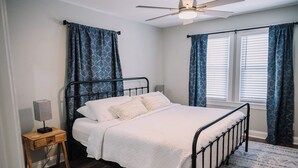Un dormitorio con cama, mesita de noche, lámpara, ventilador de techo y cortinas estampadas.