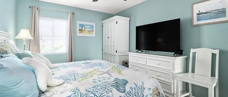 Ein Schlafzimmer mit einem Bett, einem Fernseher, einem weißen Stuhl, einem Deckenventilator und einem Fenster mit Vorhängen.