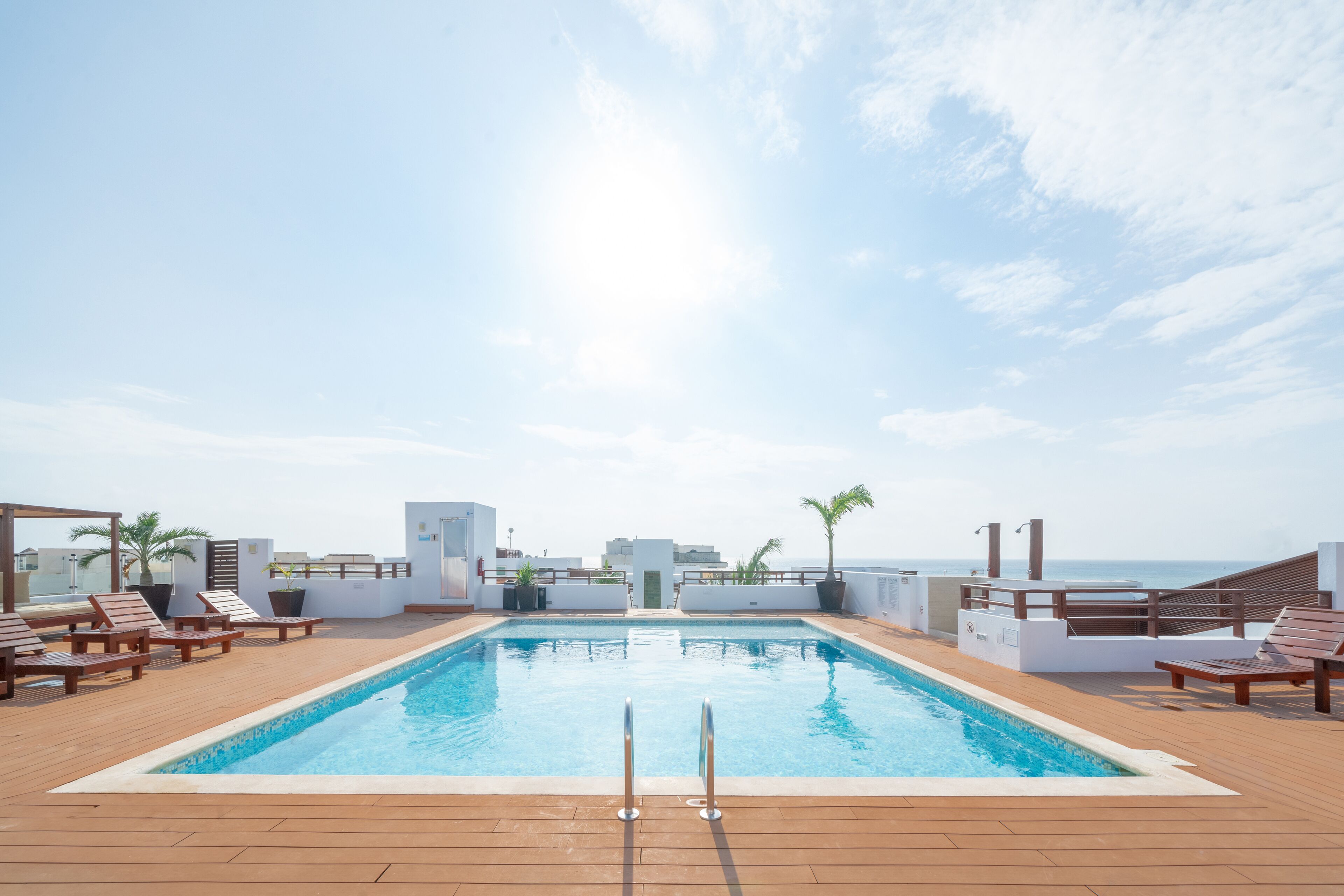 A rooftop pool with wooden deck and lounge chairs, overlooking a cityscape and the sea.