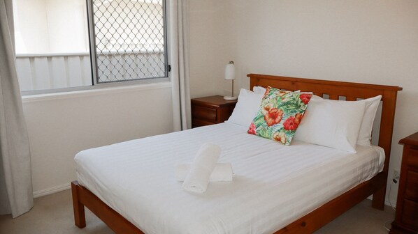 A neatly made bed with white linens and a floral pillow, a bedside lamp, and a window with a metal screen.