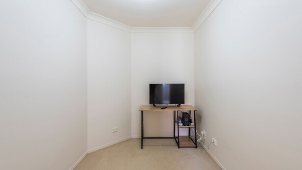 A small, empty room with white walls, a beige carpet, and a wooden TV stand with a flat-screen TV. - Oyster Cove Sunset - Stone Real Estate Yamba (Yamba)