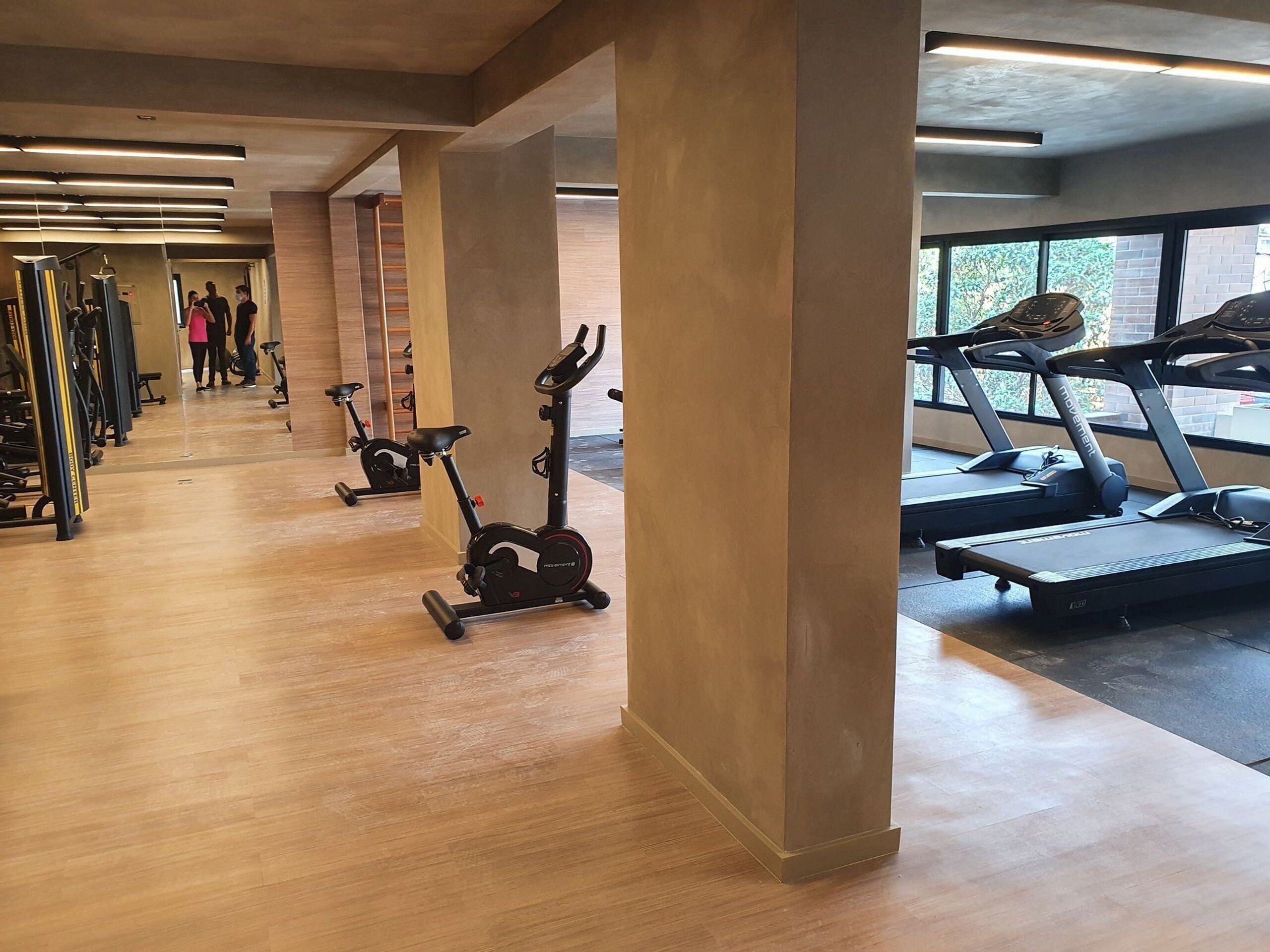 A gym with cardio machines, wooden flooring, and large windows.