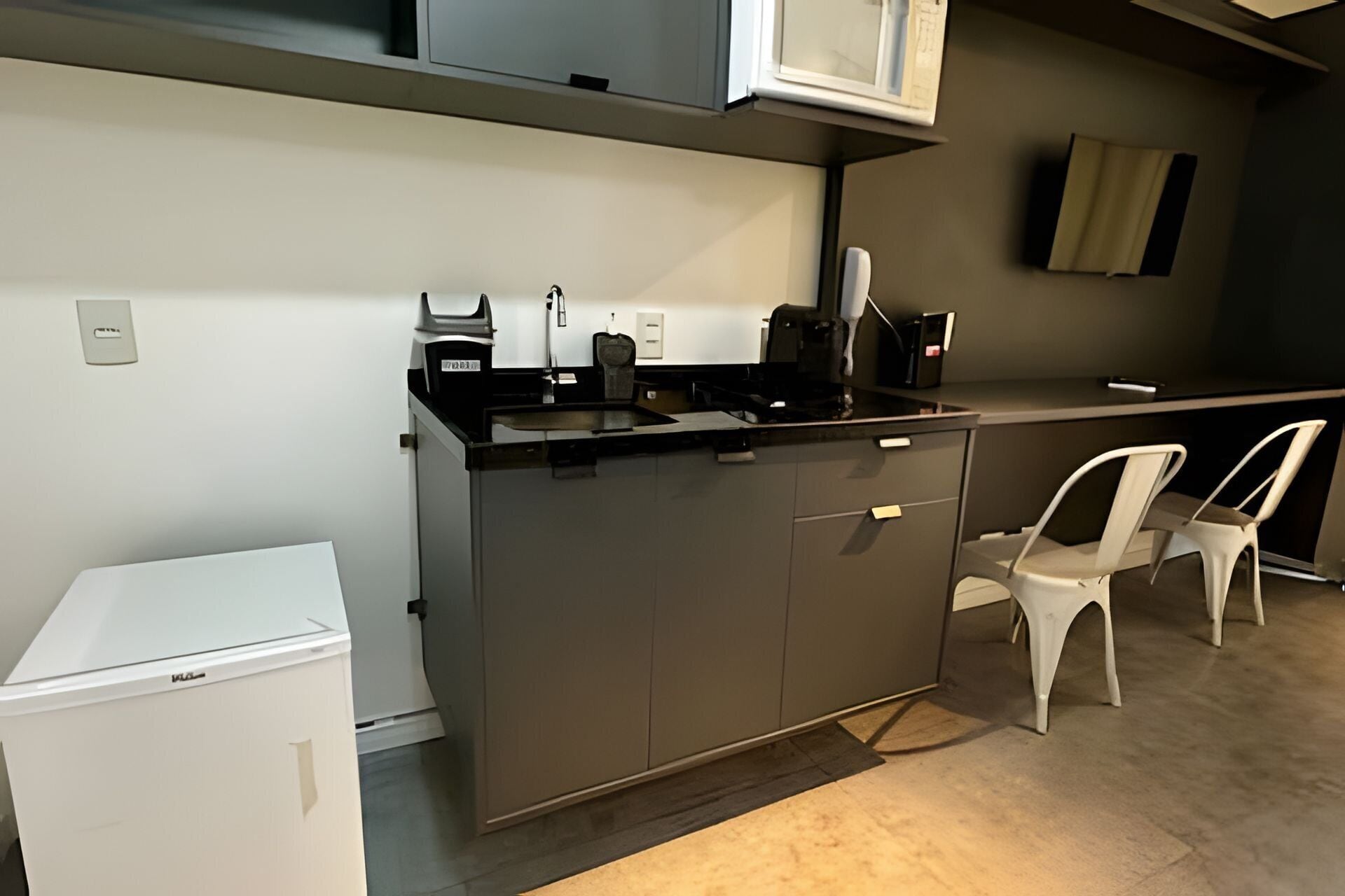 A compact kitchen with a built-in refrigerator, a coffee machine, and a microwave. There is a white chair and a small table.