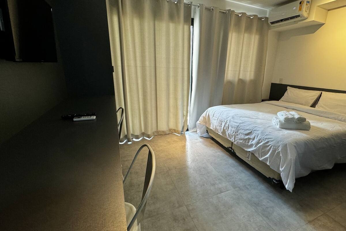 A hotel room with a bed, a chair, a remote control, and a curtain.