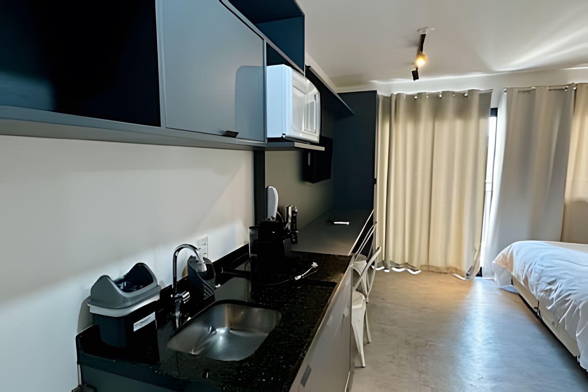 Una habitación de hotel con cama, una kitchenette pequeña con lavamanos y una ventana con cortinas.