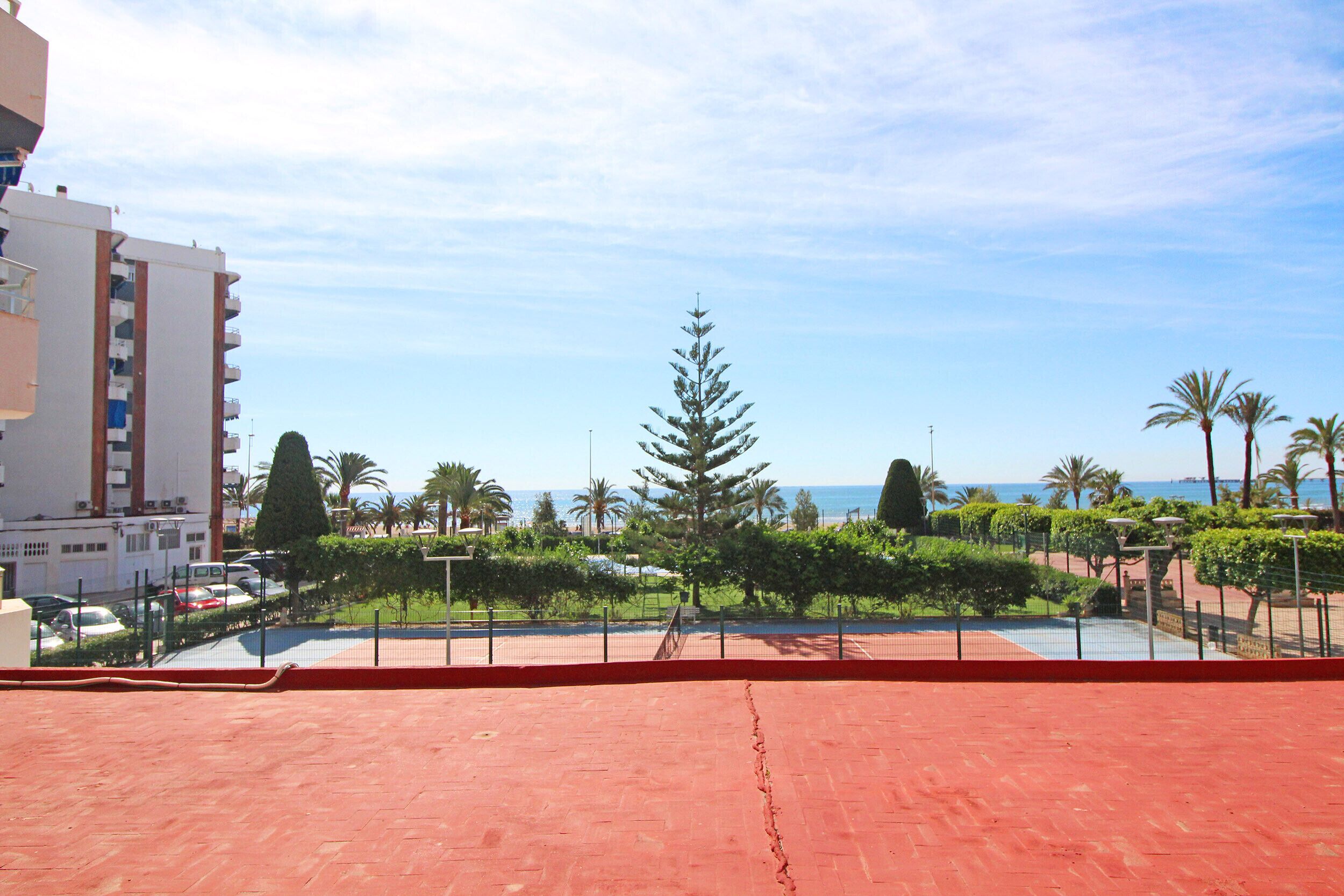A red-tiled terrace with a view of a landscaped area, palm trees, and a building in the background.