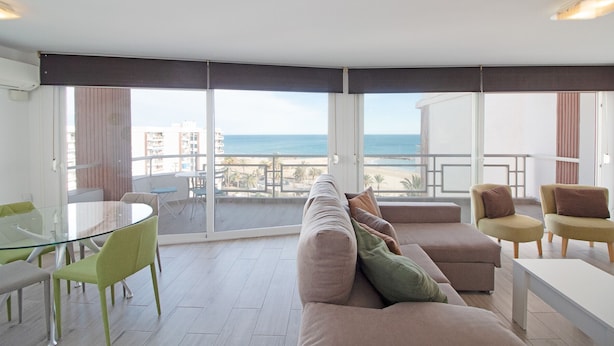 Un salon moderne avec une table en verre, des fauteuils verts et beiges, un canapé beige et une vue sur la mer et les bâtiments.